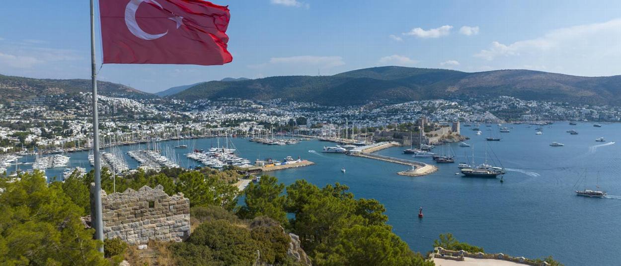 Turkish flag waving over Bodrum marina with yachts and coastlines featuring four sailboats and a historic castle in background